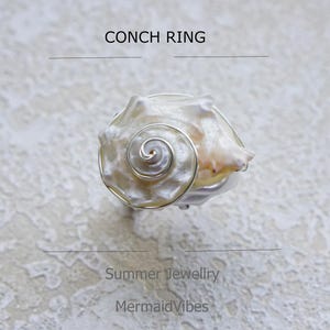 Op de afbeelding: Een conch ring met een natuurlijke zeeschelp en een zilverkleurige draadzetting. De ring heeft een spiraalvormig ontwerp en is gelabeld met "Conch Ring" en "Summer Jewellery MermaidVibes". De zeeschelp is gebroken wit en beige.
