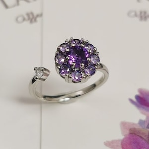 May include: A silver ring with a purple amethyst gemstone surrounded by smaller purple gemstones. The ring has a single small clear gemstone on the side.