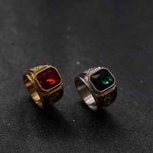 Op de afbeelding: Twee ringen met grote, vierkante edelstenen. Eén ring is goudkleurig met een rode edelsteen, de andere is zilverkleurig met een groene edelsteen. Beide hebben decoratieve details op de band, tegen een donkere achtergrond.