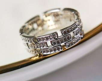 May include: A silver ring with a geometric design featuring multiple rows of small, clear cubic zirconia stones.