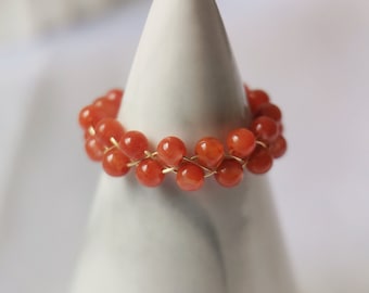 May include: A ring made of orange beads strung on a gold wire. The ring is displayed on a white cone-shaped stand.