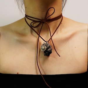 May include: A brown suede choker necklace featuring a seashell pendant. The necklace is tied in a bow at the back. The pendant is a dark, textured seashell with a spiral design.