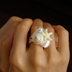 May include: A ring featuring a white spiral seashell, a small cream-coloured starfish, and a single white pearl. The band has a textured, metallic finish. The design incorporates these three distinct marine elements arranged together on the ring.