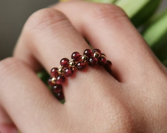 May include: A gold ring with a band of small red beads. The ring is worn on a person's finger.