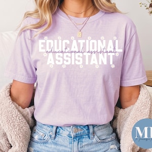 May include: A light purple short-sleeve t-shirt featuring the white text EDUCATIONAL ASSISTANT in a bold sans-serif font, with the words educational assistant in a cursive script layered over it, surrounded by small white daisy flower icons.