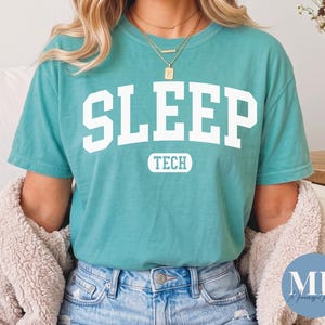 Könnte beinhalten: Ein kurzärmeliges, blaugrünes T-Shirt mit Rundhalsausschnitt, das auf der Brust das Wort SLEEP in großen weißen Blockbuchstaben zeigt, wobei das Wort TECH in Weiß in einem kleinen blaugrünen Rechteck direkt darunter steht.