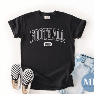 Pode incluir: Uma t-shirt preta de manga curta com o texto branco FOOTBALL numa fonte universitária arqueada sobre a palavra BRO dentro de um retângulo branco de cantos arredondados. A t-shirt é apresentada com calças de ganga azul claro e sapatilhas axadrezadas.