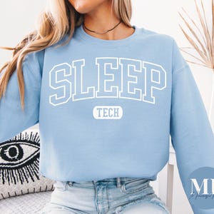 May include: A light blue crewneck sweatshirt featuring the word SLEEP in large white collegiate-style block letters across the chest, with the word TECH printed in a smaller white rounded rectangular box directly underneath the main text.