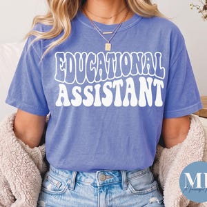 Könnte beinhalten: Ein kurzärmeliges T-Shirt in Kornblumenblau mit dem weißen Schriftzug EDUCATIONAL ASSISTANT in einer fetten, wellenförmigen Retro-Schrift auf der Brust. Das Shirt wird mit einer blauen Jeans und einer beigen, strukturierten Strickjacke getragen.