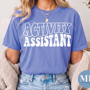Puede incluir: Una camiseta de manga corta en color azul bígaro con el texto blanco "ACTIVITY ASSISTANT" en una fuente retro, ondulada y llamativa, centrada en el pecho. La prenda tiene un cuello redondo y un corte relajado, combinada con pantalones vaqueros.