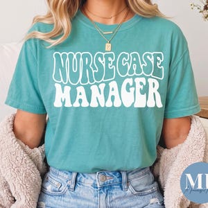 Könnte beinhalten: Ein kurzärmeliges T-Shirt in Blaugrün mit dem weißen Schriftzug NURSE CASE MANAGER in einer fetten, wellenförmigen Retro-Schriftart. Das T-Shirt wird mit einer blauen Jeans und geschichteten Goldketten getragen, darunter eine mit einem K-Anhänger.