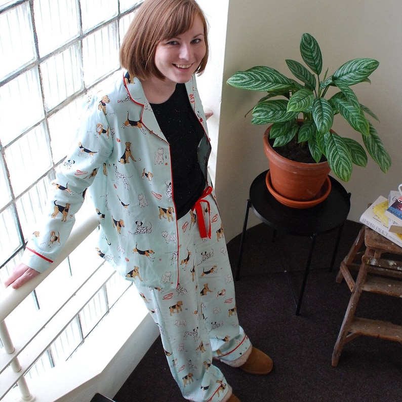 Pode incluir: Uma mulher vestindo um top e cal&ccedil;as de pijama azul claro com um padr&atilde;o de c&atilde;es. O pijama tem detalhes vermelhos e uma fita vermelha na cintura. A mulher est&aacute; em p&eacute; em frente a uma janela com uma moldura branca.