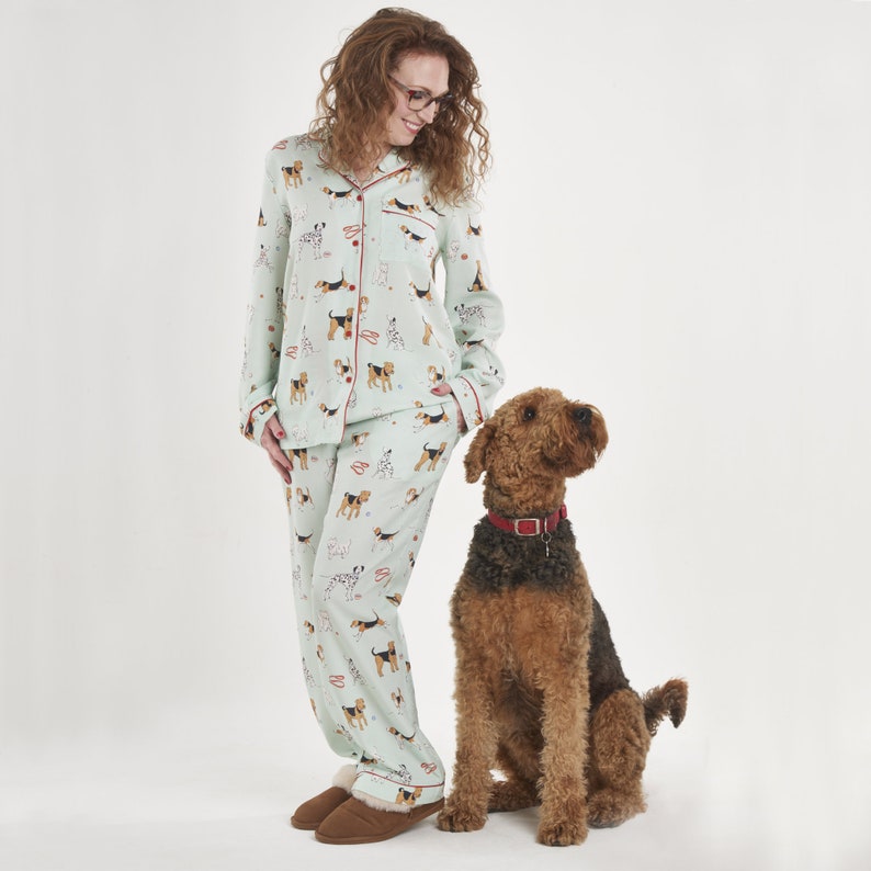 Pode incluir: Uma mulher vestindo um conjunto de pijama verde claro com estampa de cachorro. O pijama tem uma borda vermelha. Ela est&aacute; de p&eacute; ao lado de um cachorro marrom e preto.