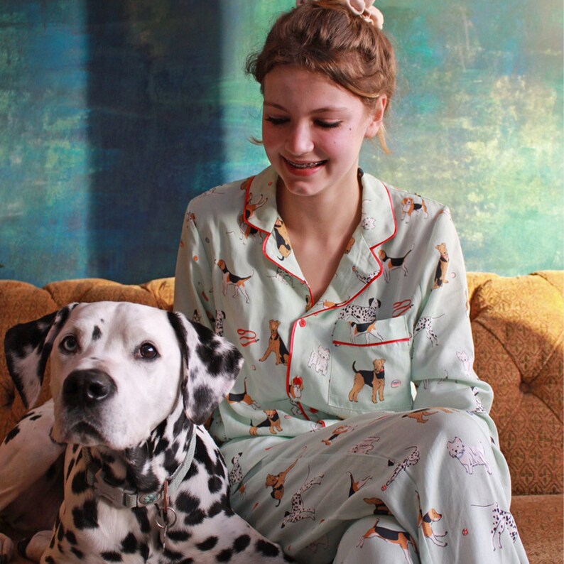 Pode incluir: Uma mulher vestindo um top de pijama azul claro com um padr&atilde;o de c&atilde;es em v&aacute;rias poses. O top tem acabamento vermelho ao redor do colarinho e punhos. Ela est&aacute; sentada em um sof&aacute; com um d&aacute;lmata ao lado dela.