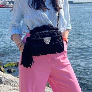 May include: A woman wearing a light blue striped shirt, pink pants, and square sunglasses. She is carrying a black woven bag with a tassel.