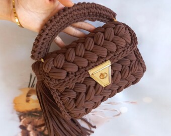 May include: A brown crocheted handbag with a gold clasp and a long tassel. The bag has a woven texture and a top handle.