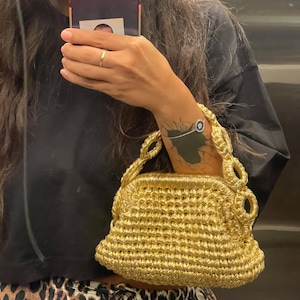 May include: A metallic gold handbag featuring a textured, crocheted construction with a structured frame closure and a braided handle. The bag has a distinct, thick woven pattern throughout its body, providing a shiny, reflective surface finish.