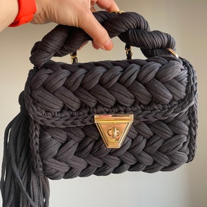 May include: A dark gray woven handbag with a braided handle and a gold clasp. The bag has a long tassel hanging from the bottom.