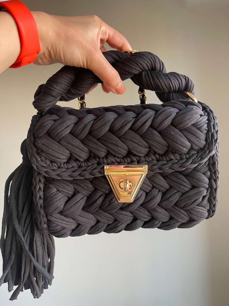 May include: A dark gray woven handbag with a braided handle and a gold clasp. The bag has a long tassel hanging from the bottom.