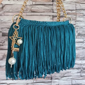 May include: A teal blue fringed handbag with a gold chain strap and a charm with a star, pearl, and bell.