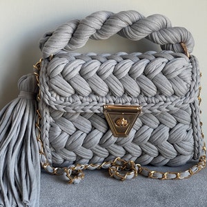 May include: A gray crocheted handbag with a braided handle and a gold chain strap. The bag has a gold clasp and a tassel.