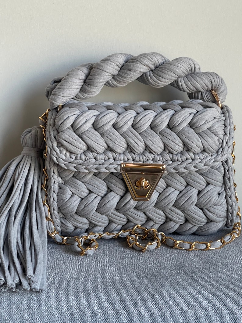 May include: A gray crocheted handbag with a braided handle and a gold chain strap. The bag has a gold clasp and a tassel.