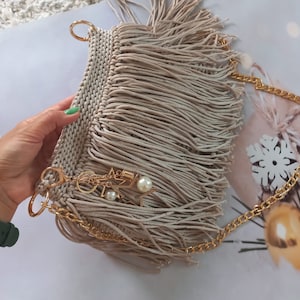 May include: A beige fringed handbag with a gold chain strap and a gold charm with a pearl. The bag is made of a woven material and has a tassel detail.
