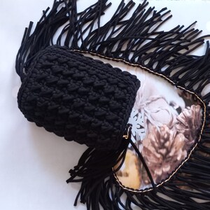 May include: Black crocheted bag with a fringe trim. The bag has a textured design and is made of thick yarn.