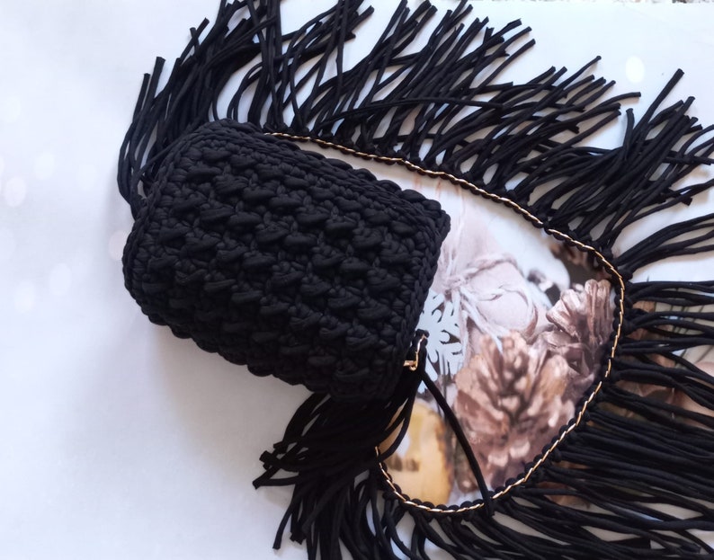 May include: Black crocheted bag with a fringe trim. The bag has a textured design and is made of thick yarn.