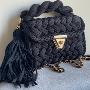 May include: A black woven handbag with a braided handle and a gold chain strap. The bag has a tassel detail and a gold clasp.