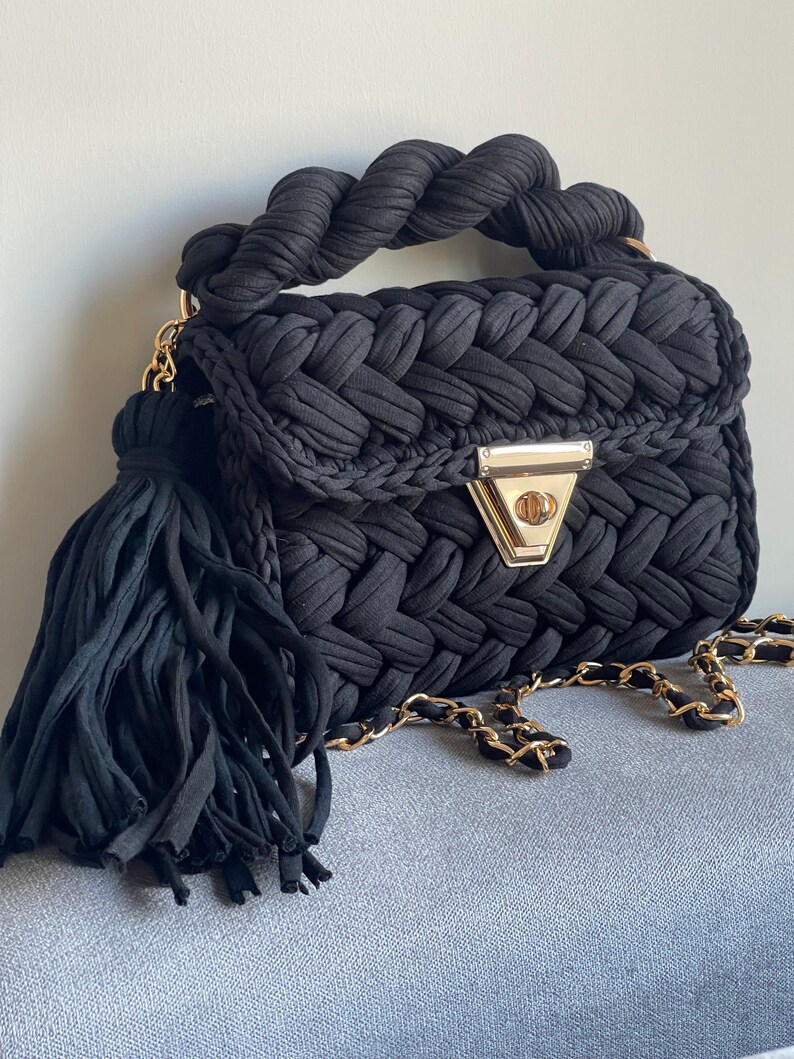 May include: A black woven handbag with a braided handle and a gold chain strap. The bag has a tassel detail and a gold clasp.