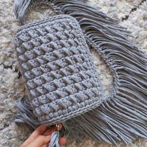 May include: A gray crocheted bag with a long fringe strap. The bag has a simple, textured design and is made with a soft, yarn-like material.