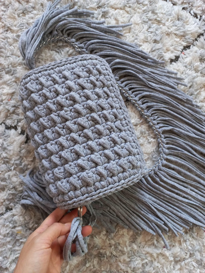 May include: A gray crocheted bag with a long fringe strap. The bag has a simple, textured design and is made with a soft, yarn-like material.
