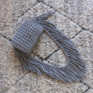 May include: A gray crocheted bag with a long fringe. The bag is lying on a white fluffy rug.