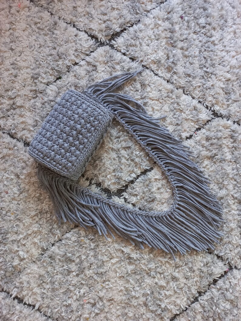 May include: A gray crocheted bag with a long fringe. The bag is lying on a white fluffy rug.