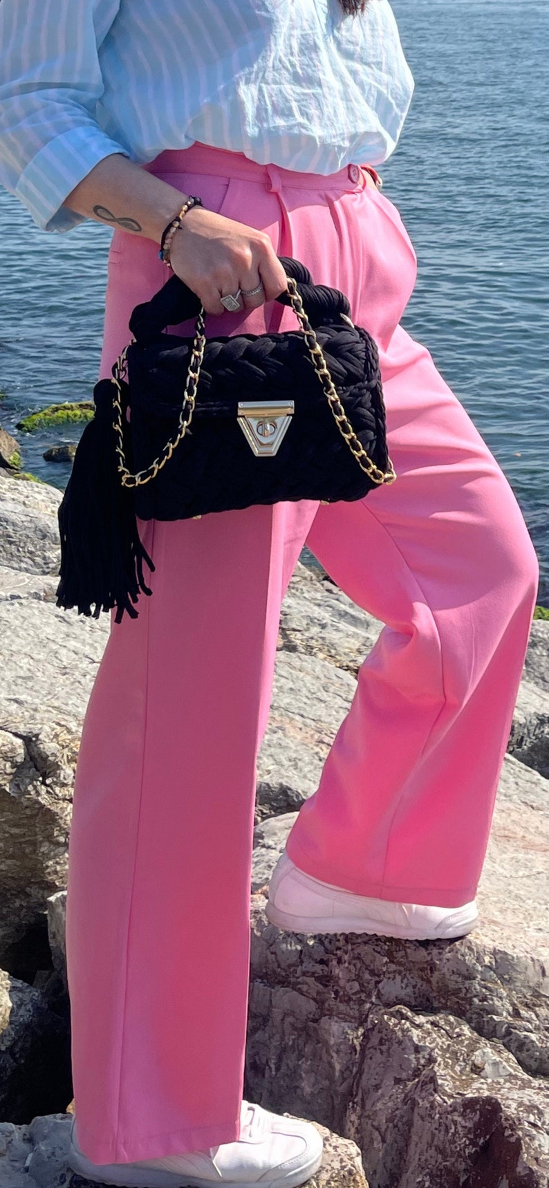 May include: A woman wearing a light blue striped shirt and pink pants is holding a black quilted handbag with a gold chain strap and a black tassel. The handbag has a gold clasp and a silver accent.