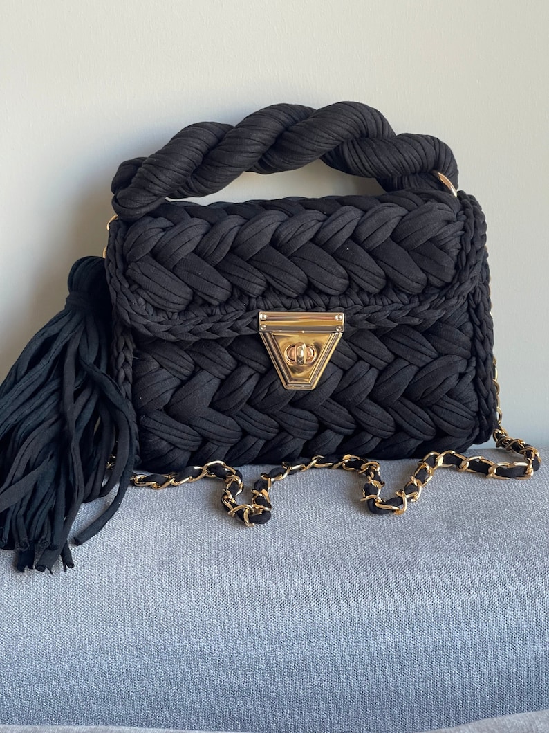 May include: A black woven handbag with a braided handle and a gold chain strap. The bag has a tassel and a gold clasp.