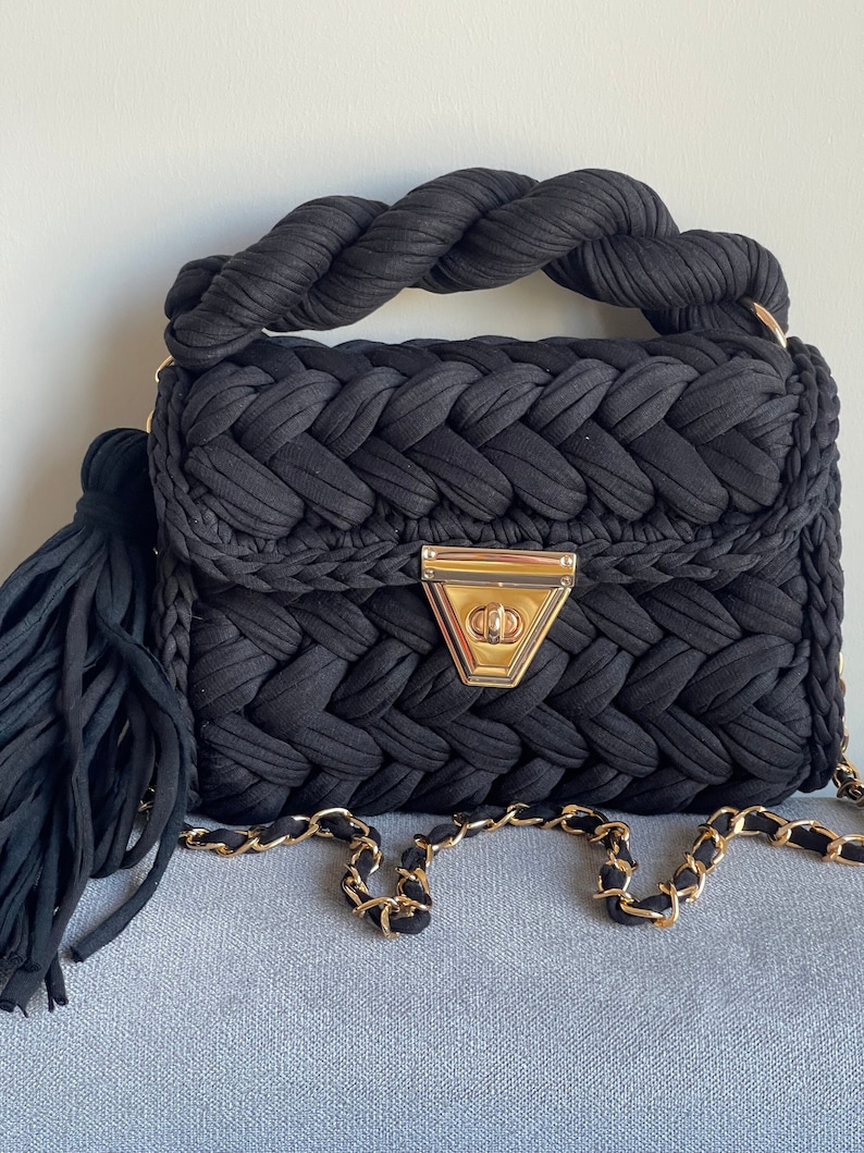 May include: A black woven handbag with a braided top handle and a gold chain strap. The bag has a gold clasp and a black tassel.