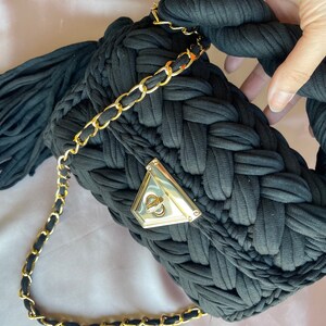 May include: Black woven fabric handbag with a gold chain strap and a gold clasp. The bag has a tassel on the side.