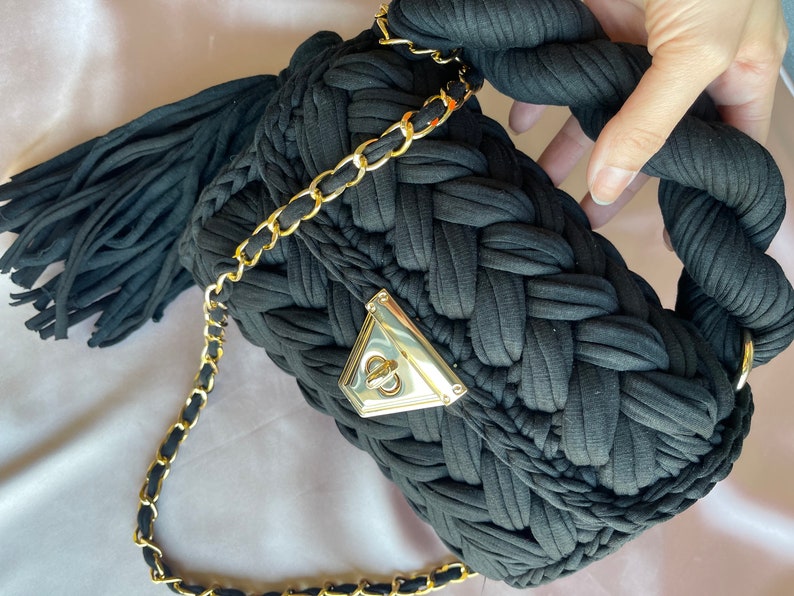 May include: Black woven fabric handbag with a gold chain strap and a gold clasp. The bag has a tassel on the side.