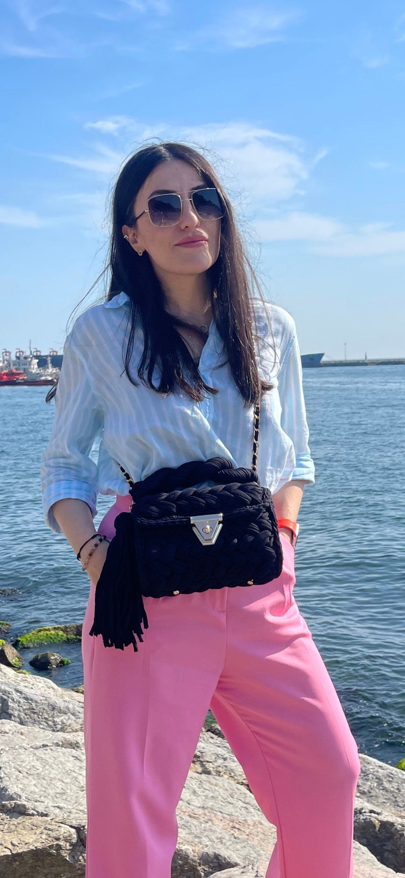 May include: A woman wearing a light blue striped shirt, pink pants, and square sunglasses. She is carrying a black woven bag with a tassel.