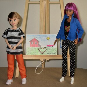 May include: Two dolls standing in front of an easel with a drawing of a house, a car, and a heart. The doll on the left is wearing a striped shirt and orange trousers. The doll on the right is wearing a blue jacket, black and white polka dot trousers, and a shirt with the text "Sweet Dream".