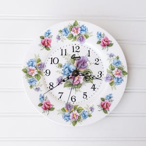 以下が含まれることがあります： ピンク、青、紫の花と緑の葉のフローラルパターンが施された白い陶器製の丸い掛け時計。時計の文字盤には1から12までの黒いアラビア数字、黒い筆記体でQuartzという文字、そして3本の黒い時計の針が表示されています。