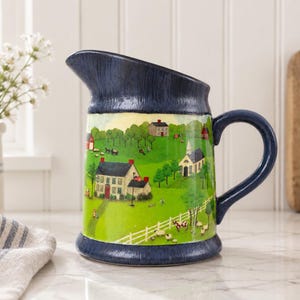 May include: A ceramic jug featuring a dark blue rim and base with a central band depicting a colourful rural landscape scene with houses, a church, green fields, trees, and a white fence. The jug has a curved handle and a wide pouring spout.