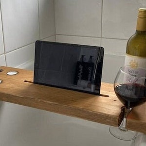 May include: A wooden bath tray with a tablet, wine glass, and bottle of wine. The tray features a slot for the tablet, a wine glass holder, and two candle holders. The wine bottle has a white label.