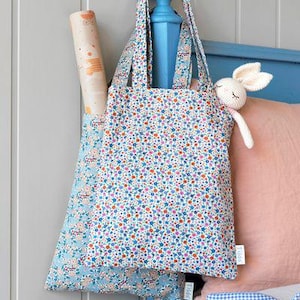 May include: Two floral print tote bags with blue and white floral patterns. The bag on top has a light blue floral pattern and the bag on the bottom has a darker blue floral pattern. The bags are hanging on a blue wooden rod.