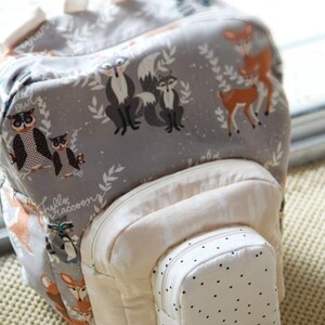 May include: A gray backpack with a woodland animal print featuring foxes, deer, and owls. The backpack has a cream-colored front pocket and a smaller, dotted pocket. The handle and straps are cream-colored.