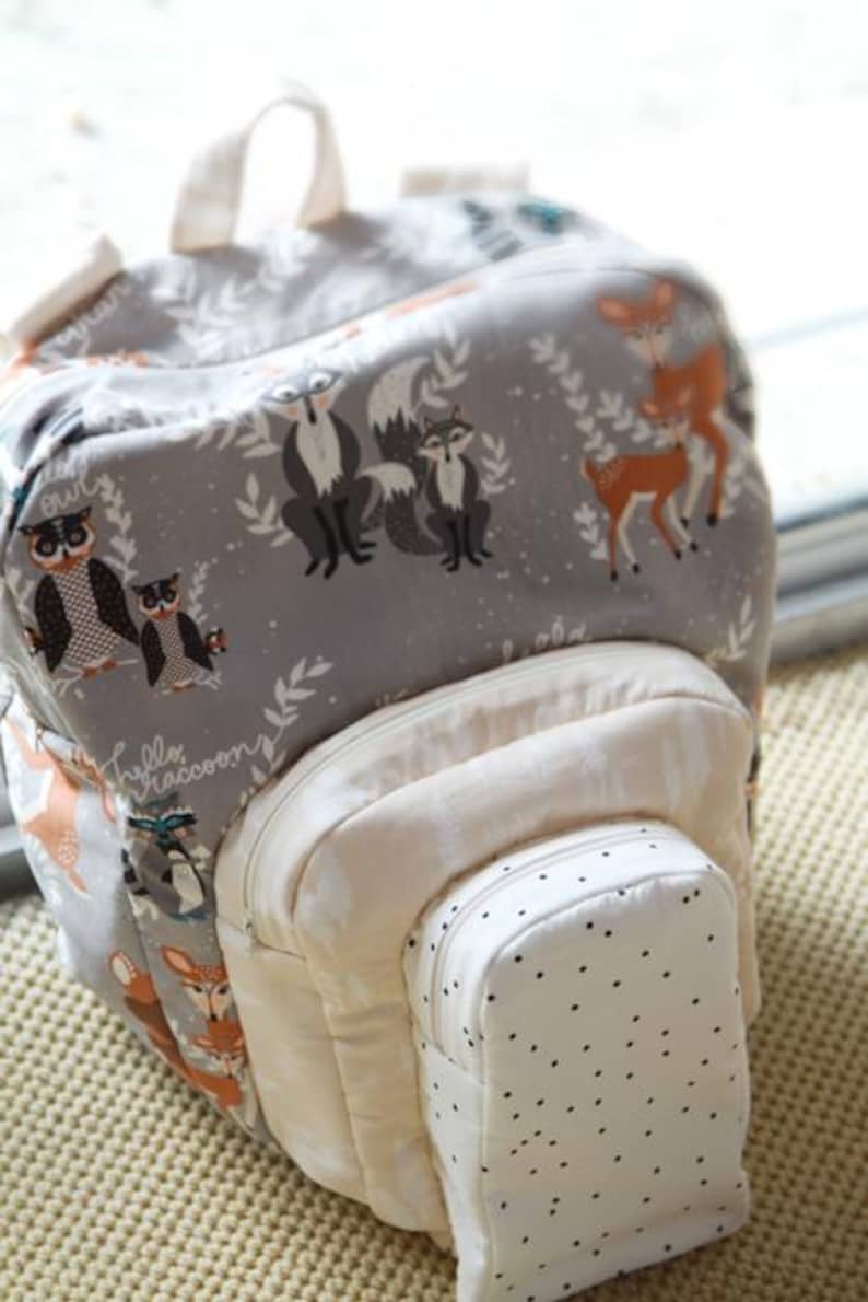 May include: A gray backpack with a woodland animal print featuring foxes, deer, and owls. The backpack has a cream-colored front pocket and a smaller, dotted pocket. The handle and straps are cream-colored.