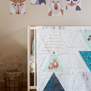May include: A baby crib with a white and blue quilt featuring triangle patterns and animal illustrations. Above the crib, framed animal portraits of a fox, raccoon, deer, bear, and owl are displayed on the wall. A woven basket with branches sits nearby.