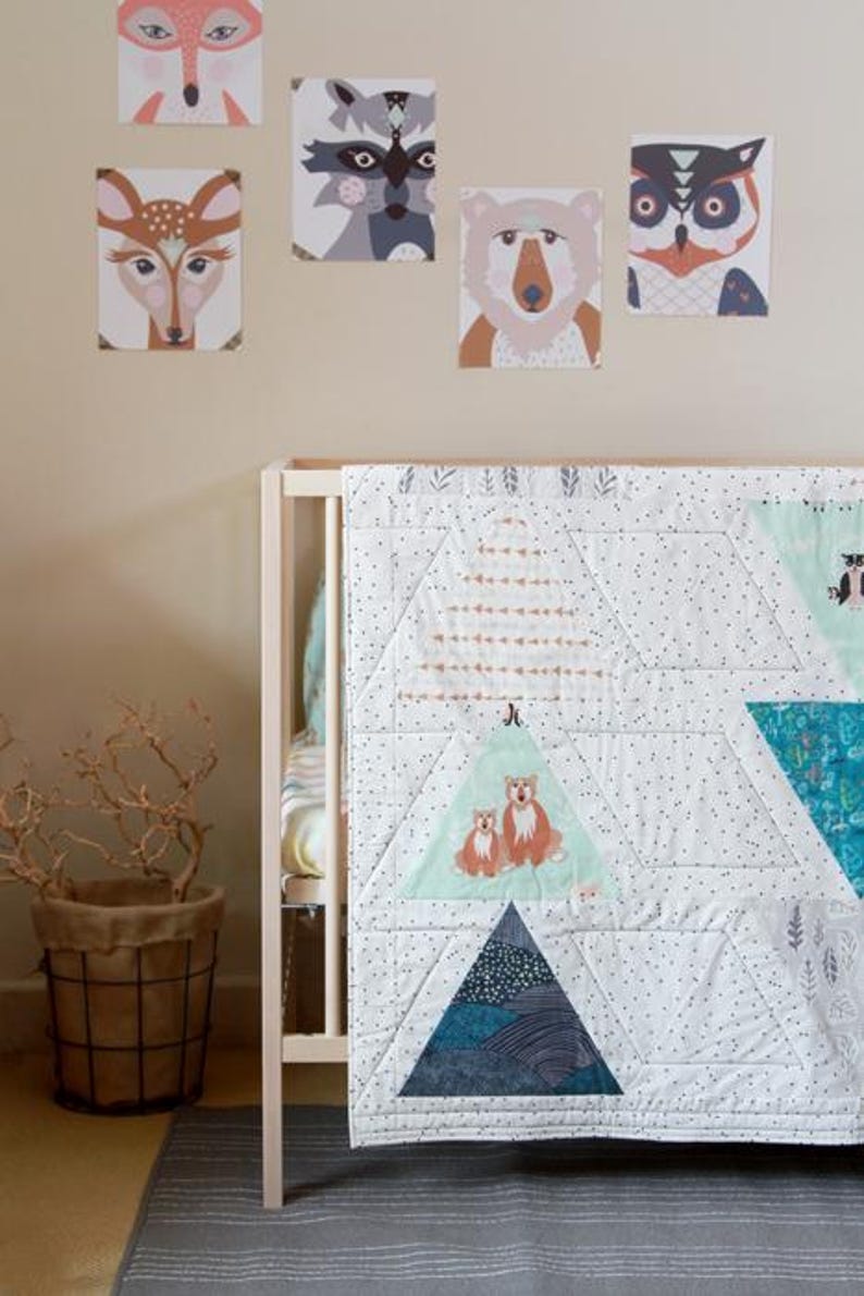May include: A baby crib with a white and blue quilt featuring triangle patterns and animal illustrations. Above the crib, framed animal portraits of a fox, raccoon, deer, bear, and owl are displayed on the wall. A woven basket with branches sits nearby.